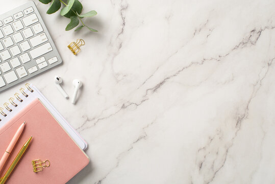 Business concept. Top view photo of workplace keyboard pink notepads wireless earbuds pens gold binder clip and eucalyptus on white marble background with empty space