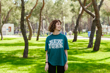Naklejka premium Young happy redhead woman wearing green t-shirt standing on green city park, smiling confident and looking away with smiles. Taking breath, meditation, planning her future.