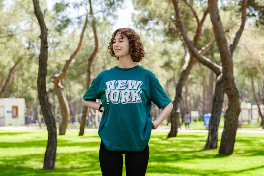 Happy young redhead woman wearing casual green tee and standing on city park, outdoors holding hands on hips and looking away with smiles, closed eyes. Smiling confidence.