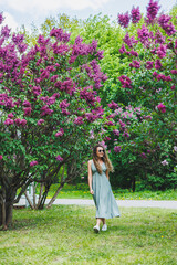 A beautiful curly woman in a summer dress runs and rejoices, she is wearing sunglasses. Against the background of a purple lilac bush