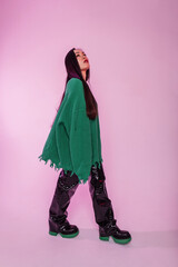 Stylish beautiful Chinese Asian model girl in fashionable casual clothes with knitted ripped green sweater in black leather pants and boots walks in the studio on a pink background