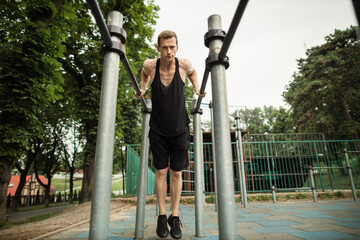 Young caucasian sporty muscular fit man doing triceps dips on parallel bars at an outdoor gym park....