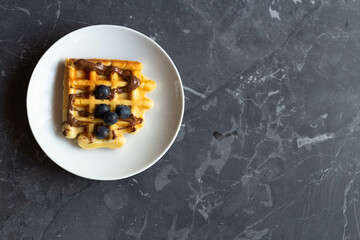Waffle with chocolate paste and blueberries on a white plate. Gray background. View from above.