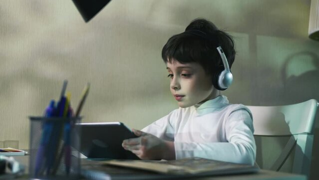 Curious Child Little school Boy Using Digital Tablet Technology Device Sitting At The Table Listening To Music Surfing Internet Play Game At Home Children Tech Addiction Concept