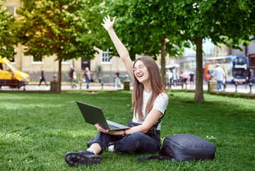 A young woman rejoices sitting on the lawn with a laptop. The girl works remotely on a laptop in the fresh air. The concept of remote work. Work in the park.