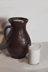 
An earthenware jug with milk and a glass of drink next to it. Photo of cow's milk, light background, soft colors