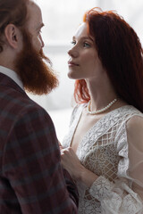 Beautiful red-hair bride in a white wedding dress and bouquet of flowers wearing pearls and a handsome groom with a red beard in a suit. Young loving couple