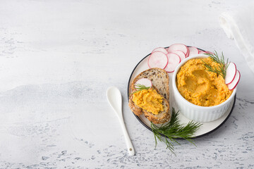 Simple egg pate or dip with carrots and fried onions in a white bowl decorated with radish slices and dill with cereal bread on a light gray background