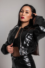 Fashionable portrait of a stylish Asian girl with makeup in a fashion black leather jacket with bra and pants on a gray background