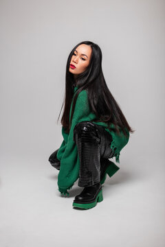 Fashionable beautiful young woman model with long hair in a fashionable green sweater with leather pants and leather boots sits and poses on a gray background