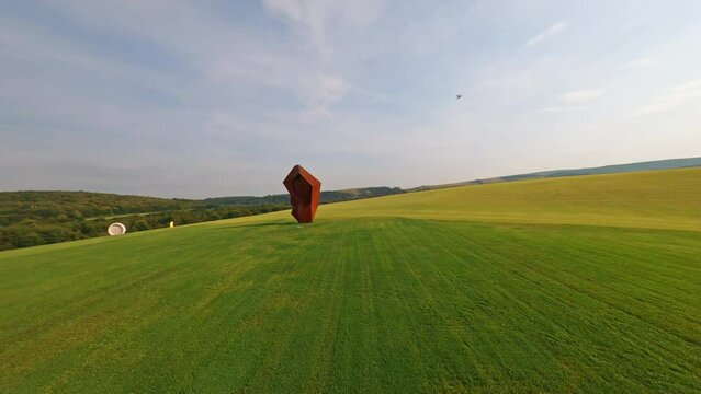 Open space. FPV fast flight near cottage town. Speed motion. Green field with burgundy and gray sculptures. Modern Art. Eclecticism. Aerial follow view. Park of future. Creative idea of creator.