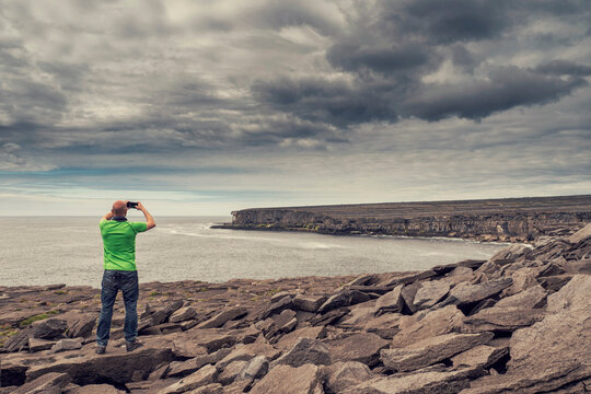 Male tourist on a rough stone surface of a cliff taking picture on phone. Ocean and dramatic cloudy sky in the background. Aran island, Ireland. Stunning Irish landscape. Travel and tourism concept.