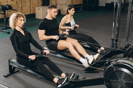 Young Man And Beautiful Women Working Out With Rowing Machine At Crossfit Gym.