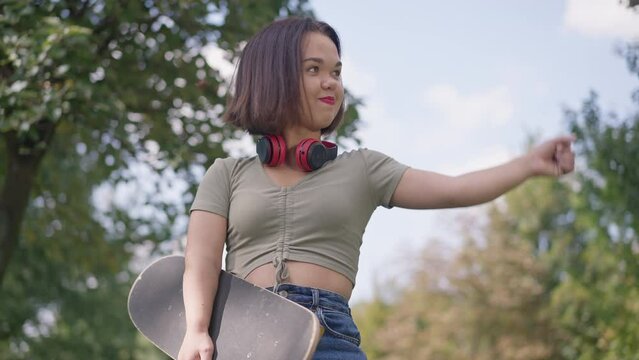 Medium shot portrait of positive little woman with skateboard gesturing strength looking at camera smiling. Happy Caucasian young person with dwarfism posing in slow motion in summer park