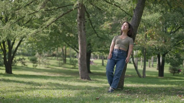 Wide shot summer park forest on sunny day with Caucasian young little woman leaning on tree trunk looking away standing on the right. Happy relaxed person with dwarfism dreaming outdoors