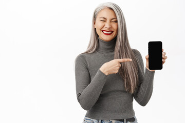 Fototapeta premium Portrait of happy asian middle aged woman laughing and smiling, pointing at smth funny on smartphone screen, showing mobile phone app or website, white background