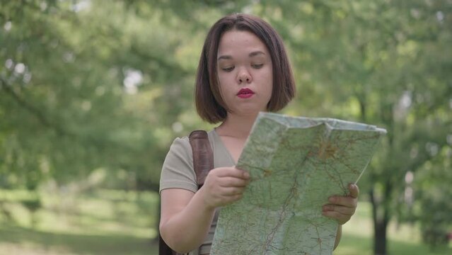 Portrait of young concentrated little woman examining paper map standing in spring summer forest outdoors. Confident Caucasian tourist with dwarfism travelling on sunny day. Slow motion