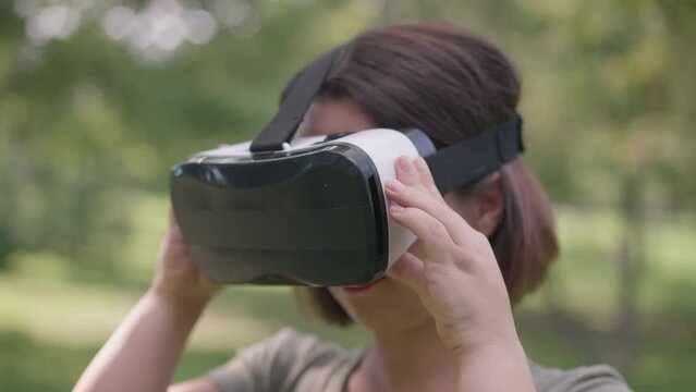 Close-up young woman with dwarfism putting on VR headset in slow motion standing in spring summer park outdoors. Little person enjoying gaming on weekend