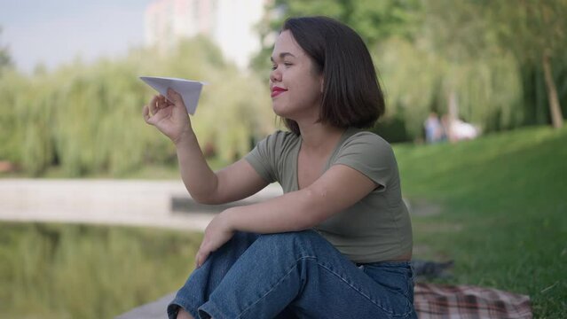 Portrait of happy carefree Caucasian little woman with paper plane sitting on lake shore in summer park. Smiling young person with dwarfism enjoying leisure outdoors on weekend