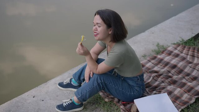 Wide shot happy relaxed positive little woman blowing soap bubbles having fun resting on lake shore in park. High angle view portrait of Caucasian young person with dwarfism enjoying leisure outdoors