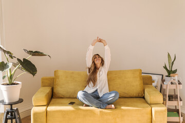 Light-skinned adult woman pulls her arms up while sitting on comfortable sofa in light room. Pretty...
