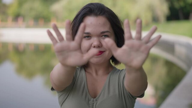 Front view positive little person with dwarfism stretching hands looking at camera smiling in slow motion. Portrait of confident happy Caucasian woman posing outdoors in spring summer park