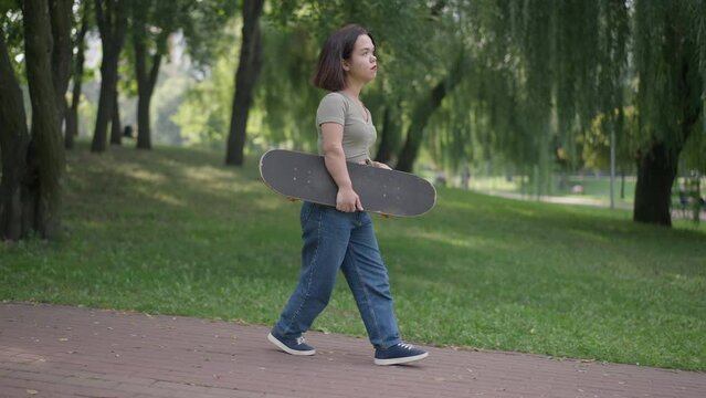Side view young Caucasian beautiful woman with dwarfism strolling with skateboard in park. Tracking shot of fit little person walking in slow motion outdoors on weekend