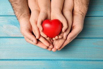 Parents and kid holding red heart in hands at light blue wooden table, top view