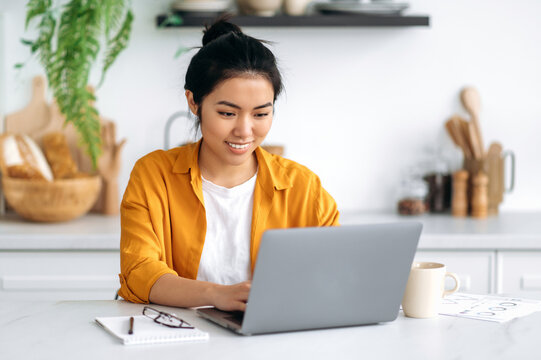 Cute attractive positive Chinese brunette girl, in stylish casual clothes, freelancer, student or designer, working on a project in a laptop, sitting at home in the kitchen, smiling