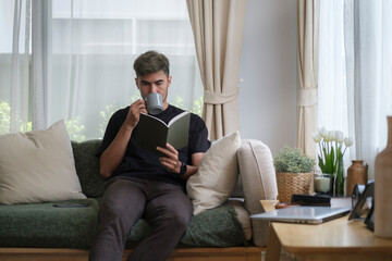 Positive young man reading book and drinking coffee while sitting on sofa at home.