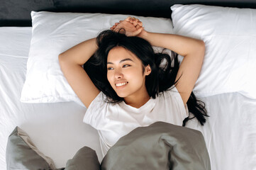 Top view of a lovely Asian brunette girl in a white t-shirt, lying under the covers in bed at home...