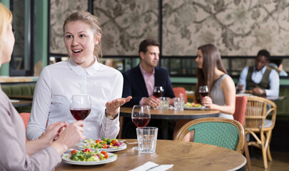 Happy attractive girl emotionally discussing with female friend while dining in cozy restaurant