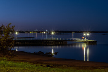 A tranquil view of a pier and city lights in the distance casting reflection on the calm sea.