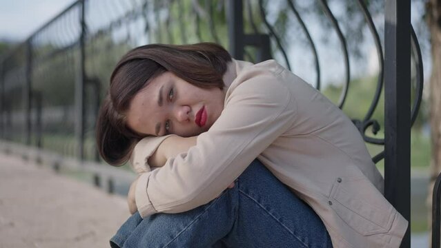 Side view portrait of depressed Caucasian young beautiful woman with dwarfism hugging knees looking away thinking. Upset frustrated little person procrastinating on bridge in summer spring park