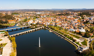 Fototapeta premium Picturesque aerial view of Mirandela city with residential houses with brownish tiled roofs, bridges crossing Tua river and water fountain on spring day, Braganca, Portugal