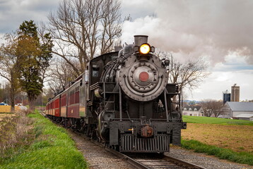 Obraz premium An Antique Steam Passenger Train Approaching on a Single Track Blowing Smoke on a Cloudy Day