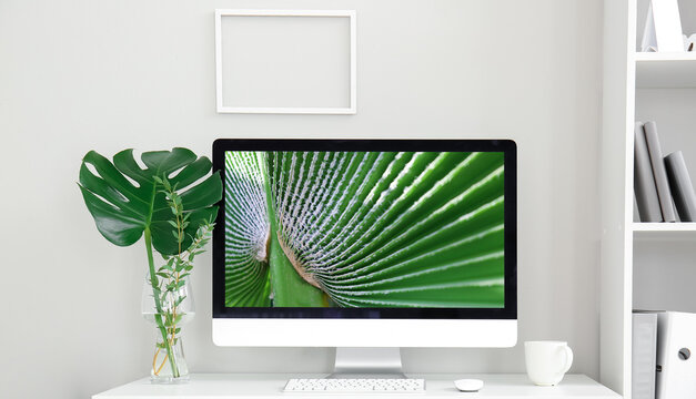 Workplace with modern computer and tropical leaf in vase near light wall