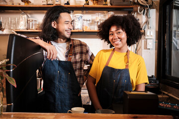 Two cafe business startup partners and friends, African American female, and Thai male baristas talk and cheerful smile together at counter bar of coffee shop, happy service job, and SME entrepreneur.