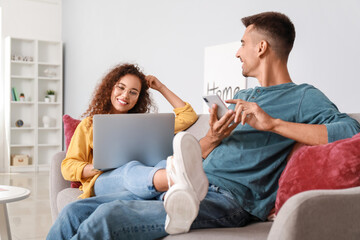 Young woman with laptop and her husband using mobile phone on sofa at home - obrazy, fototapety, plakaty