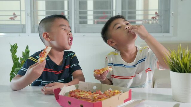 Happy Asian two little boy 5 years sitting eating huge pizza together on table at home, children eating unhealthy food, Excited funny kids eating pizza, Preschool mealtime with friends