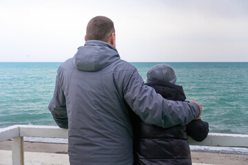  Adult son hugs his old mother, holding on to the railing and looking at the cold winter sea. Standing on the beach in winter clothes. selective focus.