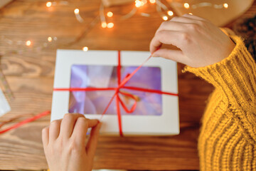 Woman in orange sweater pulls ribbon on gift box. First-person view of girl's hand opening Christmas surprise. Wooden table with garland. New Year's holiday mood.