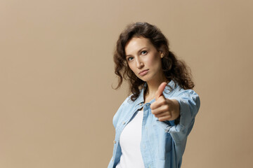 Serious confident curly beautiful female in jeans casual shirt show thumb up gesture Like at camera posing isolated on over beige pastel background. People Lifestyle emotions concept. Copy space