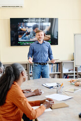 Vertical shot of mature Caucasian man standing at desk in front of multi-ethnic students teaching...