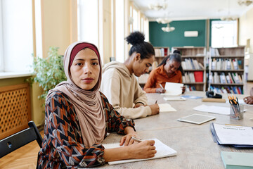 Young woman wearing hijab sitting at table with her classmates looking at camera during English...