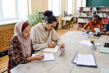 Group of multi-ethnic immigrant students sitting at table watching educational videos on digital...