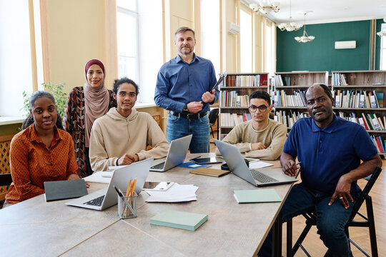 Group portrait of modern English language teacher and multi-ethnic immigrant students having class in library looking at camera