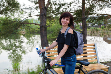 A happy female tourist with a backpack rides a bicycle in a park along the river.