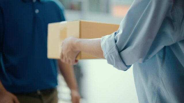 Close up parcel in hand of blue uniform delivery man to customer front of the house with good service from shopping online. Courier man delivering a cardboard box postal package to destination.