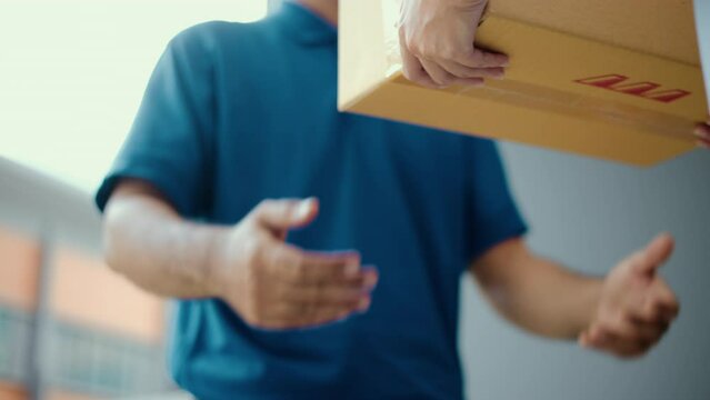 Close up parcel in hand of blue uniform delivery man to customer front of the house from shopping online with good service. Courier man delivering a cardboard box postal package to destination.